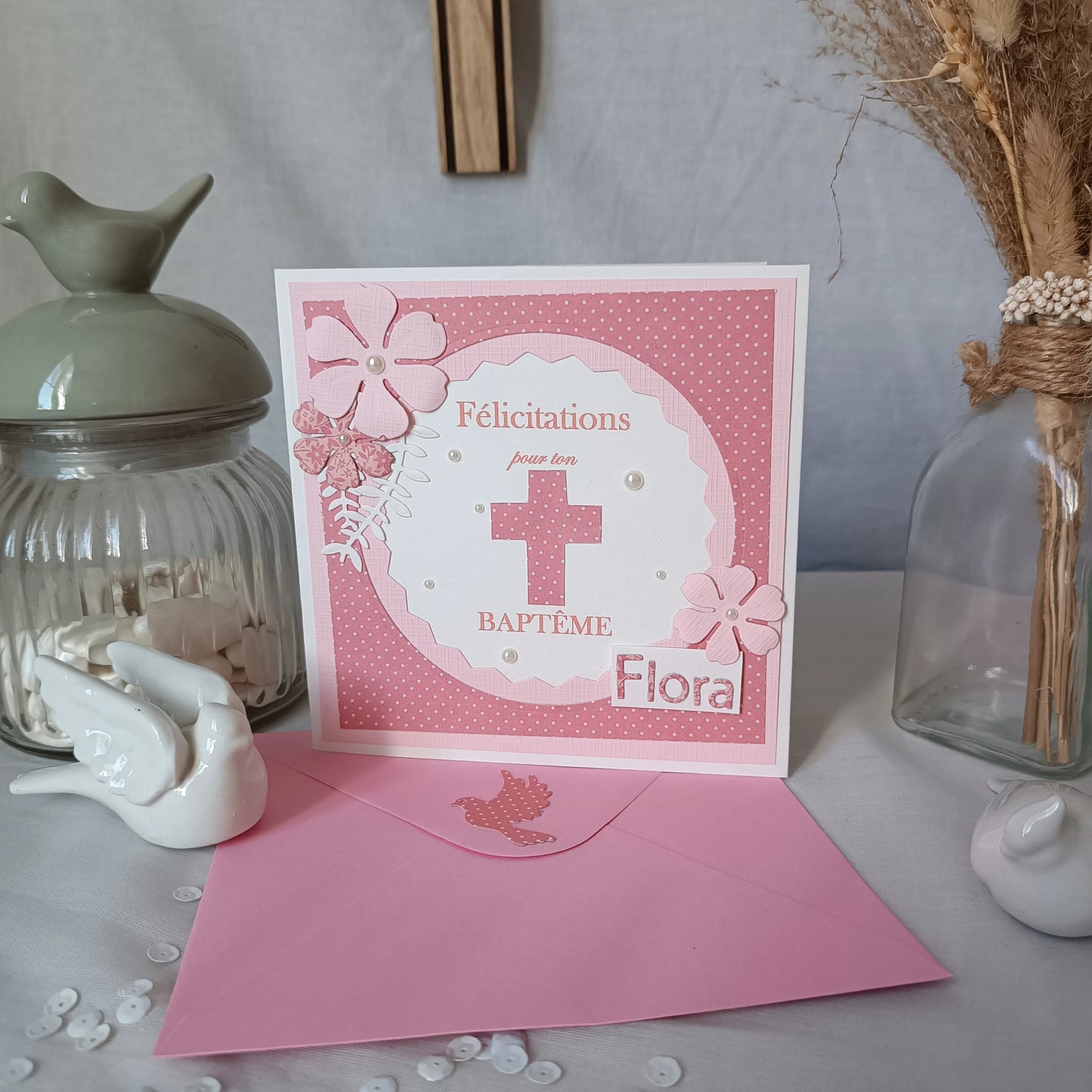 Carte de baptême rose personnalisable debout sur son enveloppe, entourée d'un pot décoratif et de fleurs séchées