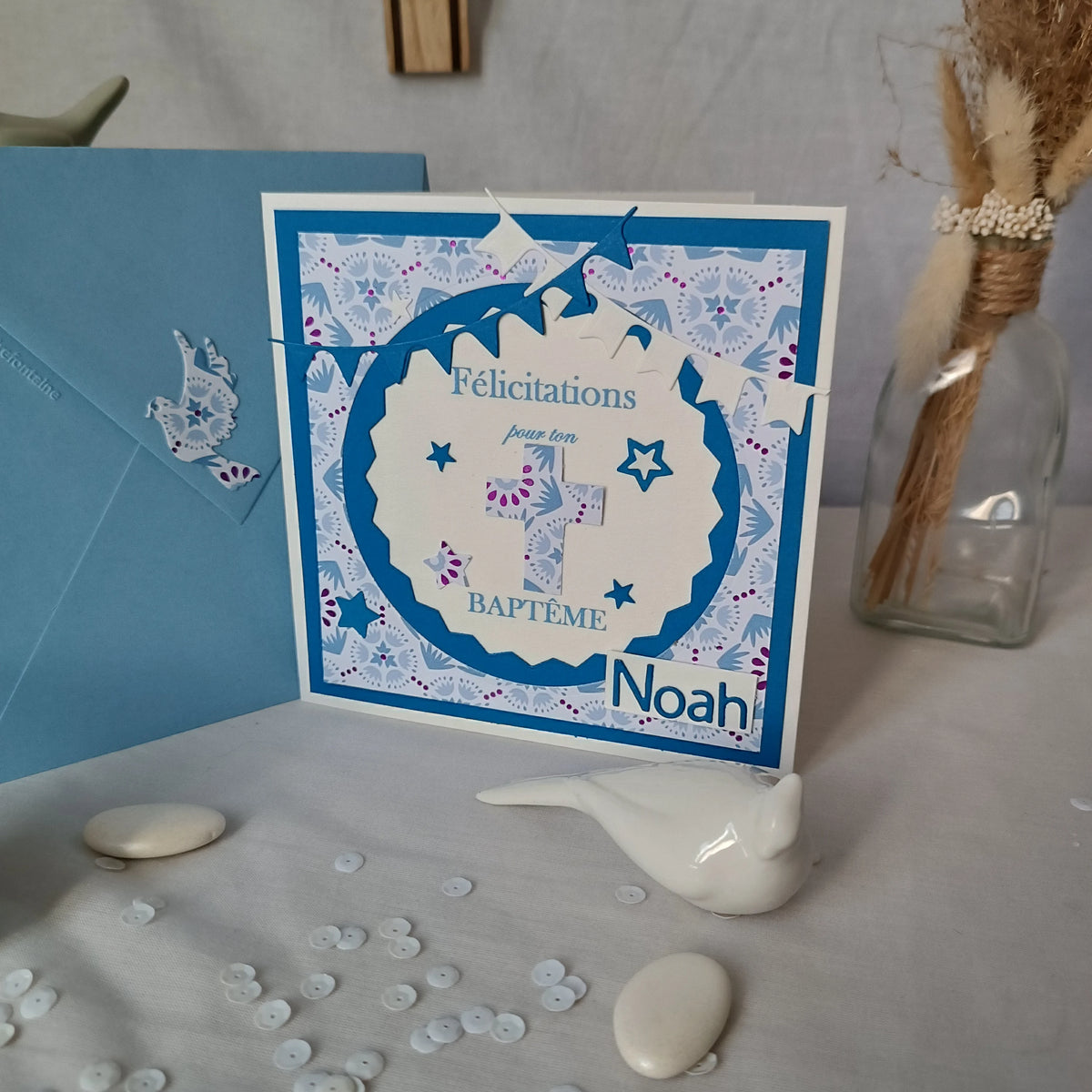 Carte de baptême bleue personnalisable posée devant un pot décoratif à couvercle oiseau