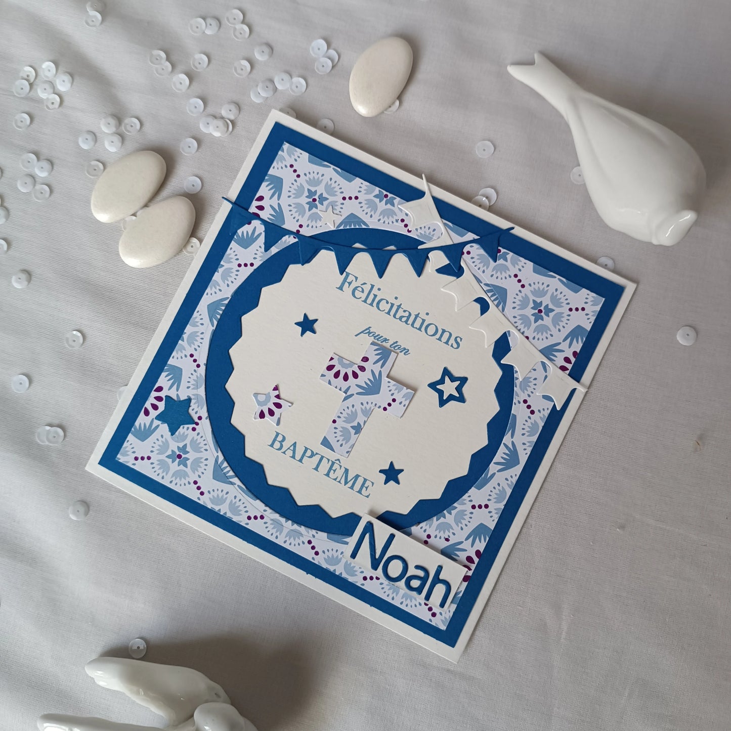 Gros plan sur la carte de baptême bleue personnalisable, avec sequins et galet blanc en premier plan