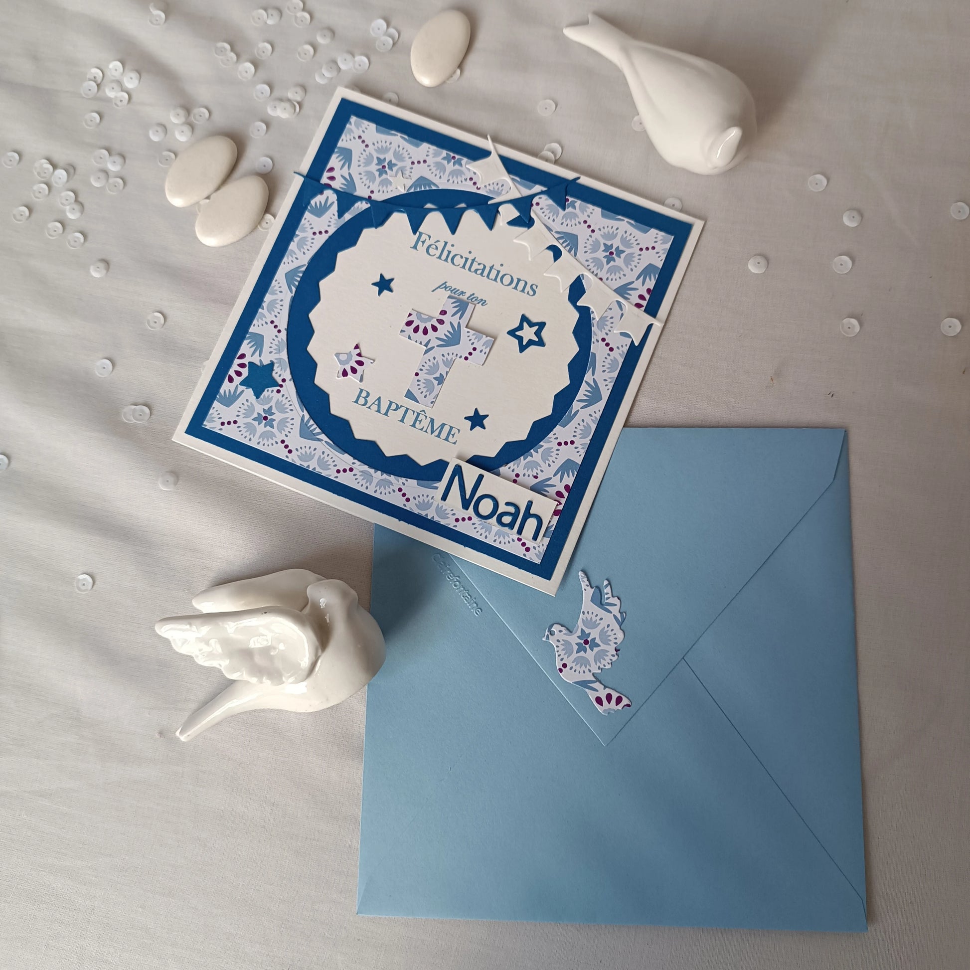 Vue plongeante de la carte de baptême bleue personnalisable posée à plat, entourée de dragées et d'oiseaux en céramique