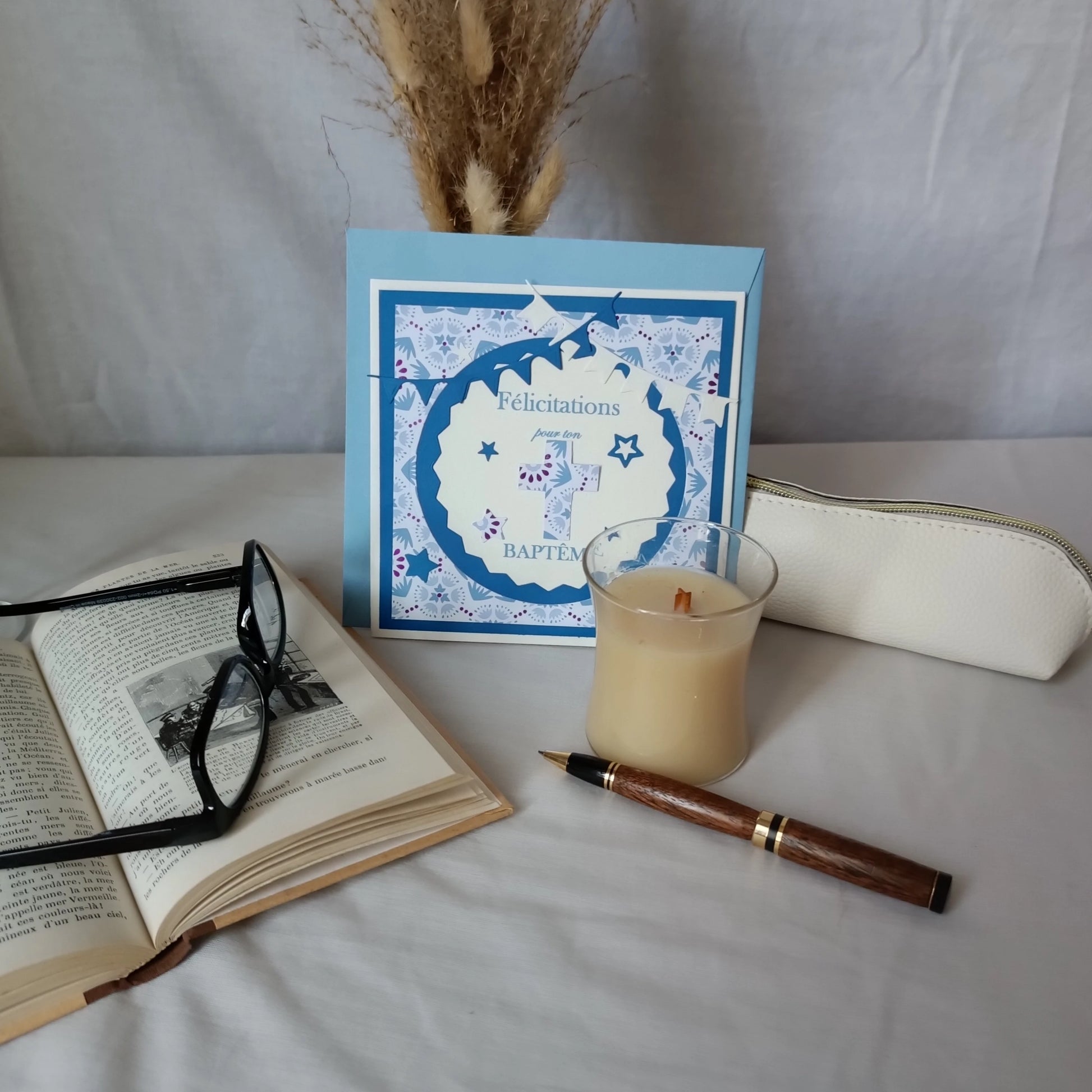 Carte de baptême bleue personnalisable debout sur son enveloppe, avec livre ancien, lunettes et bougie