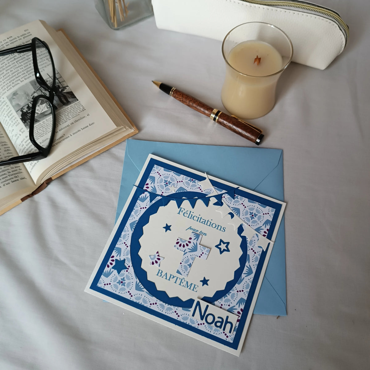 Vue plongeante de la carte de baptême bleue personnalisable sur son enveloppe, entourée d'un livre ancien et d'une bougie
