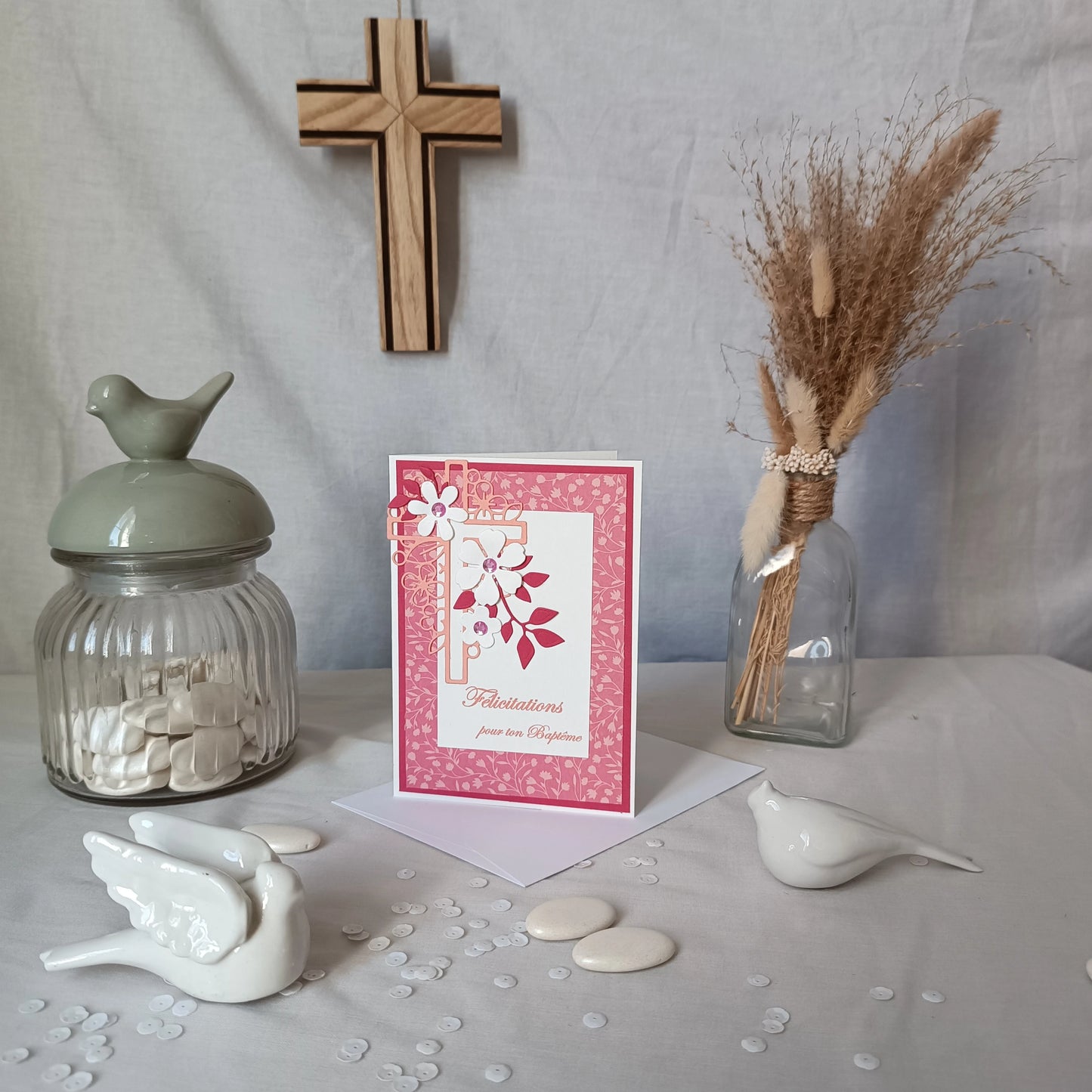 Carte de baptême rose debout sur son enveloppe, avec croix en bois, oiseau en céramique et fleurs séchées en arrière-plan
