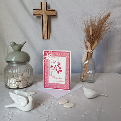 Carte de baptême rose debout sur son enveloppe, avec croix en bois, oiseau en céramique et fleurs séchées en arrière-plan