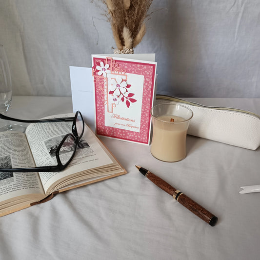 Carte de baptême rose debout devant un vase de fleurs séchées, avec livre ancien, lunettes et bougie