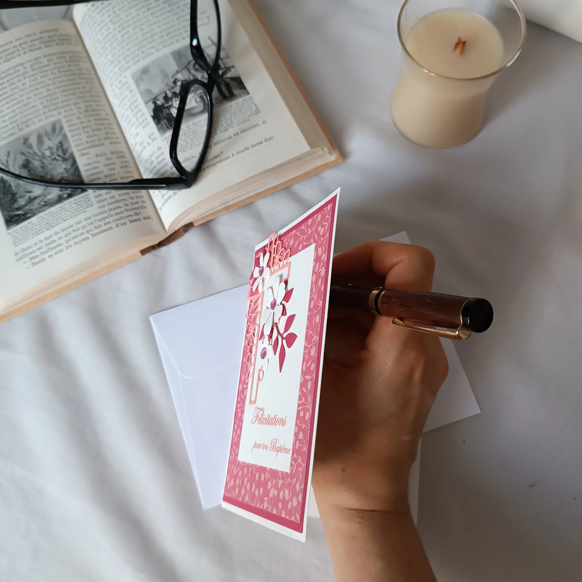 Main tenant un stylo au-dessus d'une carte de baptême rose posée sur son enveloppe blanche, décor avec livre ancien et bougie