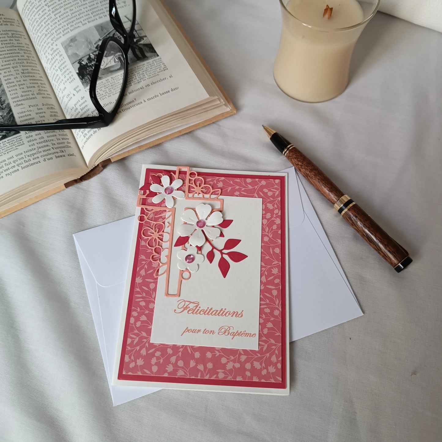 Carte de baptême rose posée sur son enveloppe, entourée d'un stylo, d'un livre ancien ouvert et d'une bougie