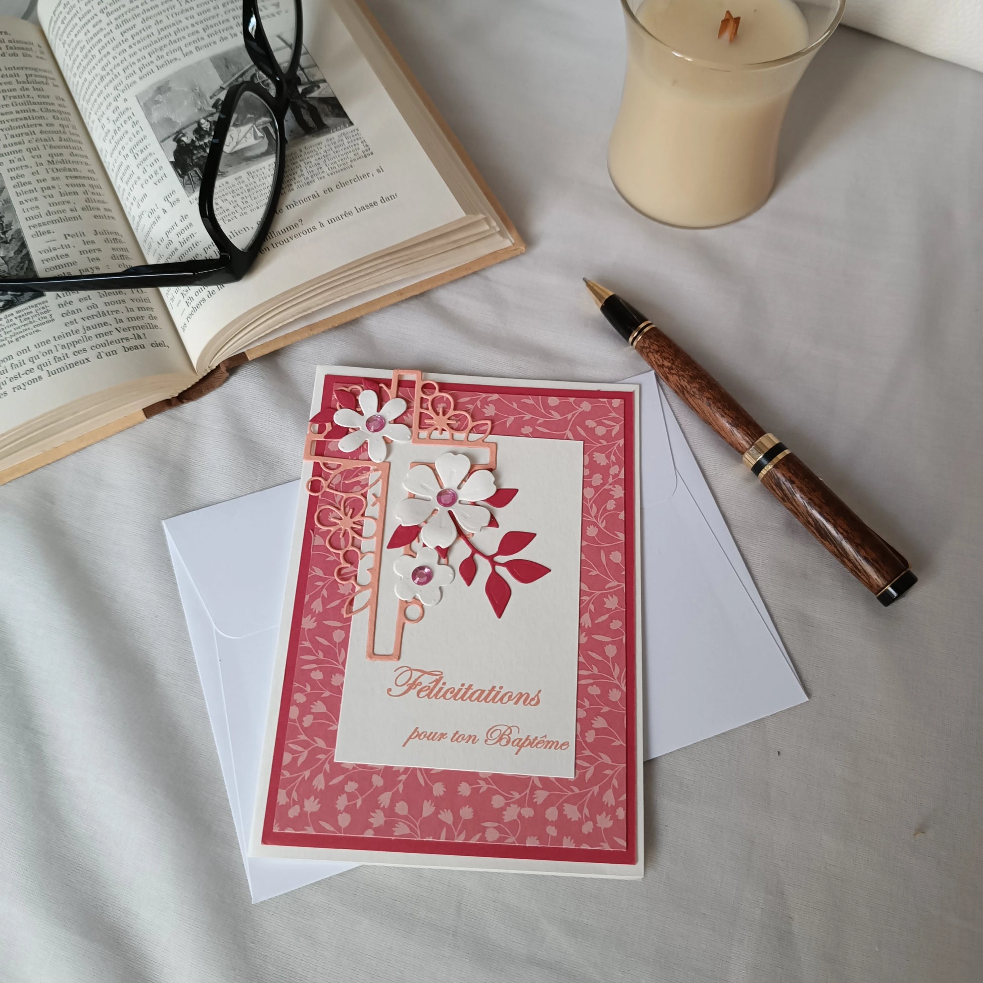 Carte de baptême rose posée sur son enveloppe, entourée d'un stylo, d'un livre ancien ouvert et d'une bougie
