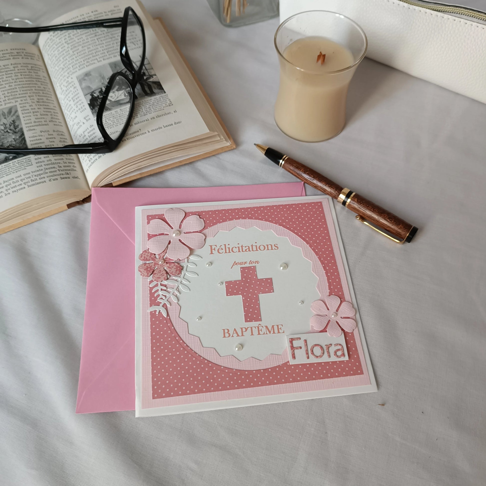 Carte de baptême rose personnalisable posée sur son enveloppe, entourée d'un livre ancien, de lunettes et d'un stylo