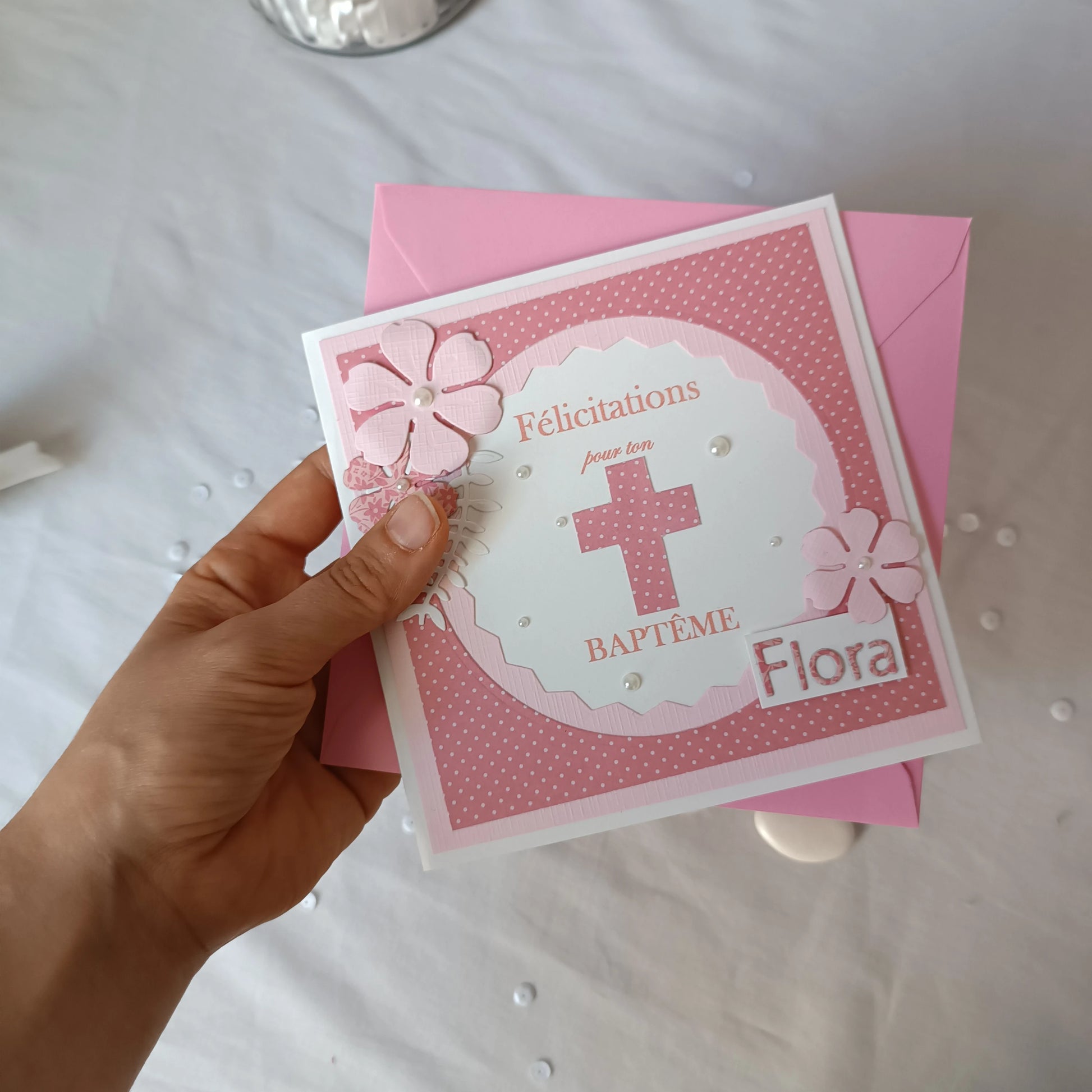 Main tenant la carte de baptême rose personnalisable devant son enveloppe assortie