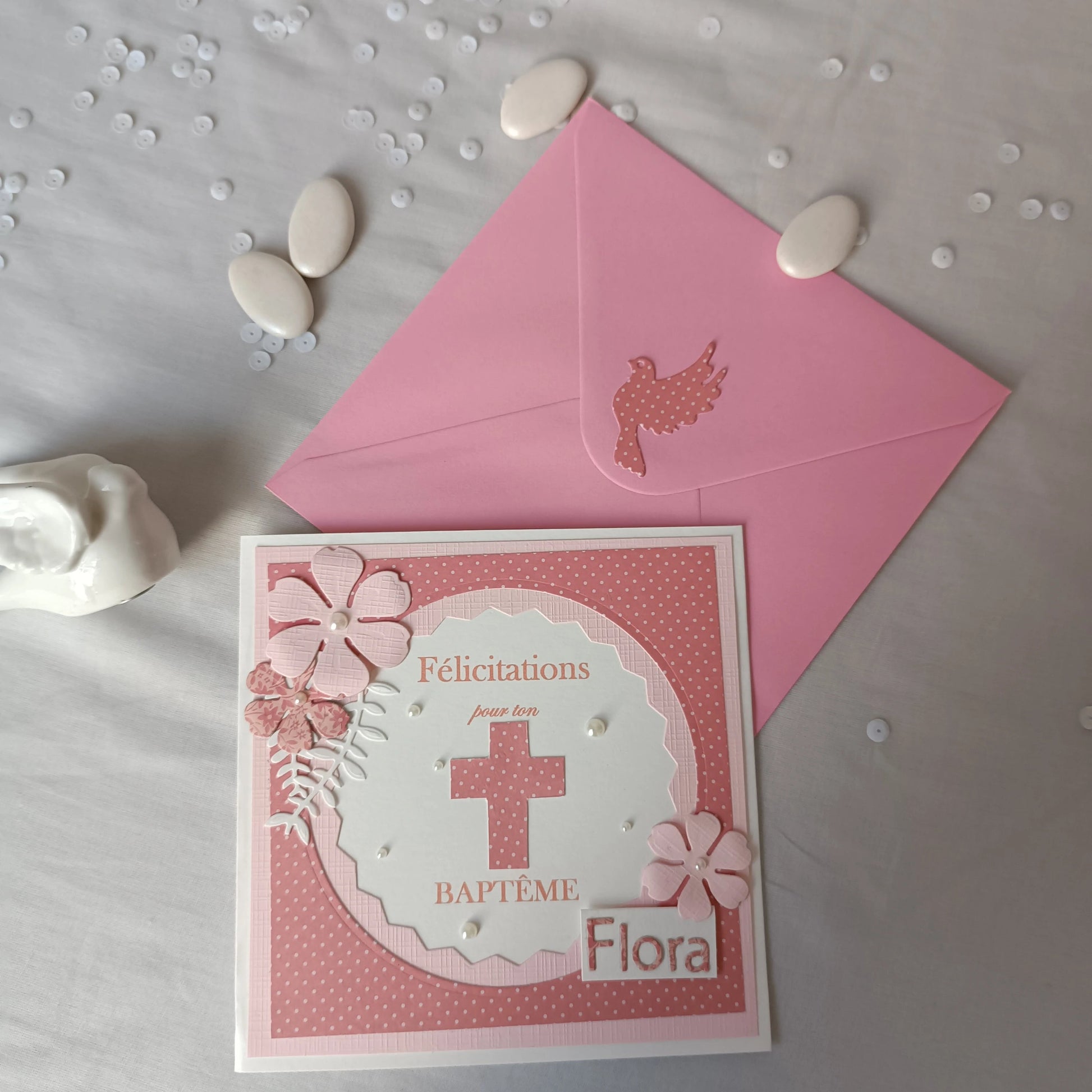 Carte de baptême rose personnalisable posée en diagonale sur son enveloppe, avec dragées et sequins