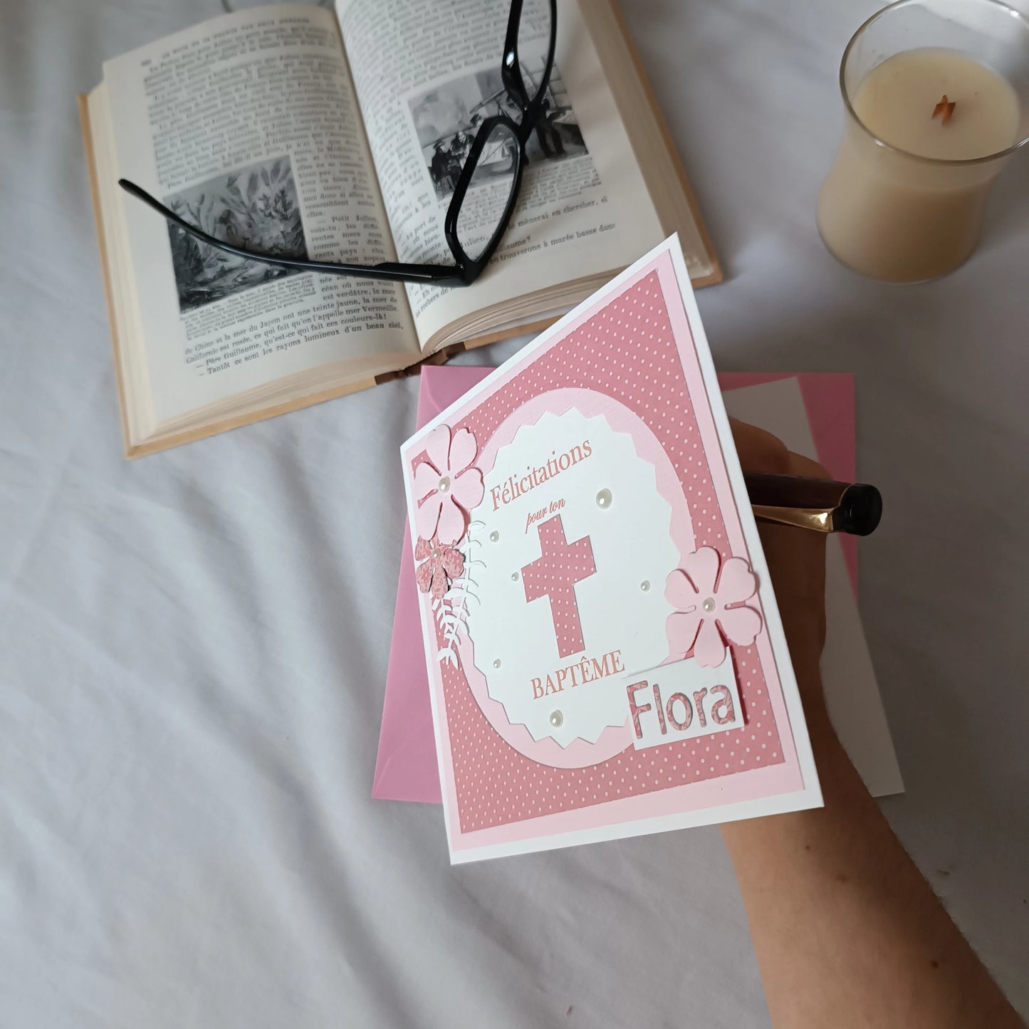 Carte de baptême rose personnalisable tenue en main au-dessus de son enveloppe, avec livre ancien et bougie