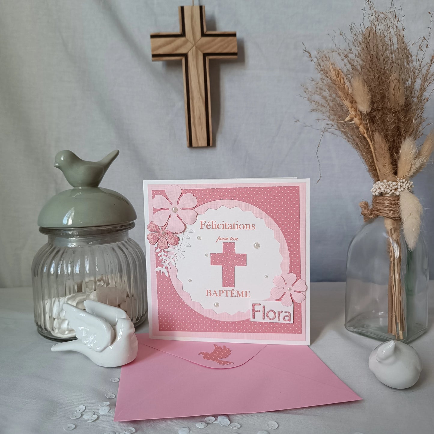 Carte de baptême rose personnalisable, debout sur son enveloppe rose, avec croix en bois et pot décoratif en arrière-plan