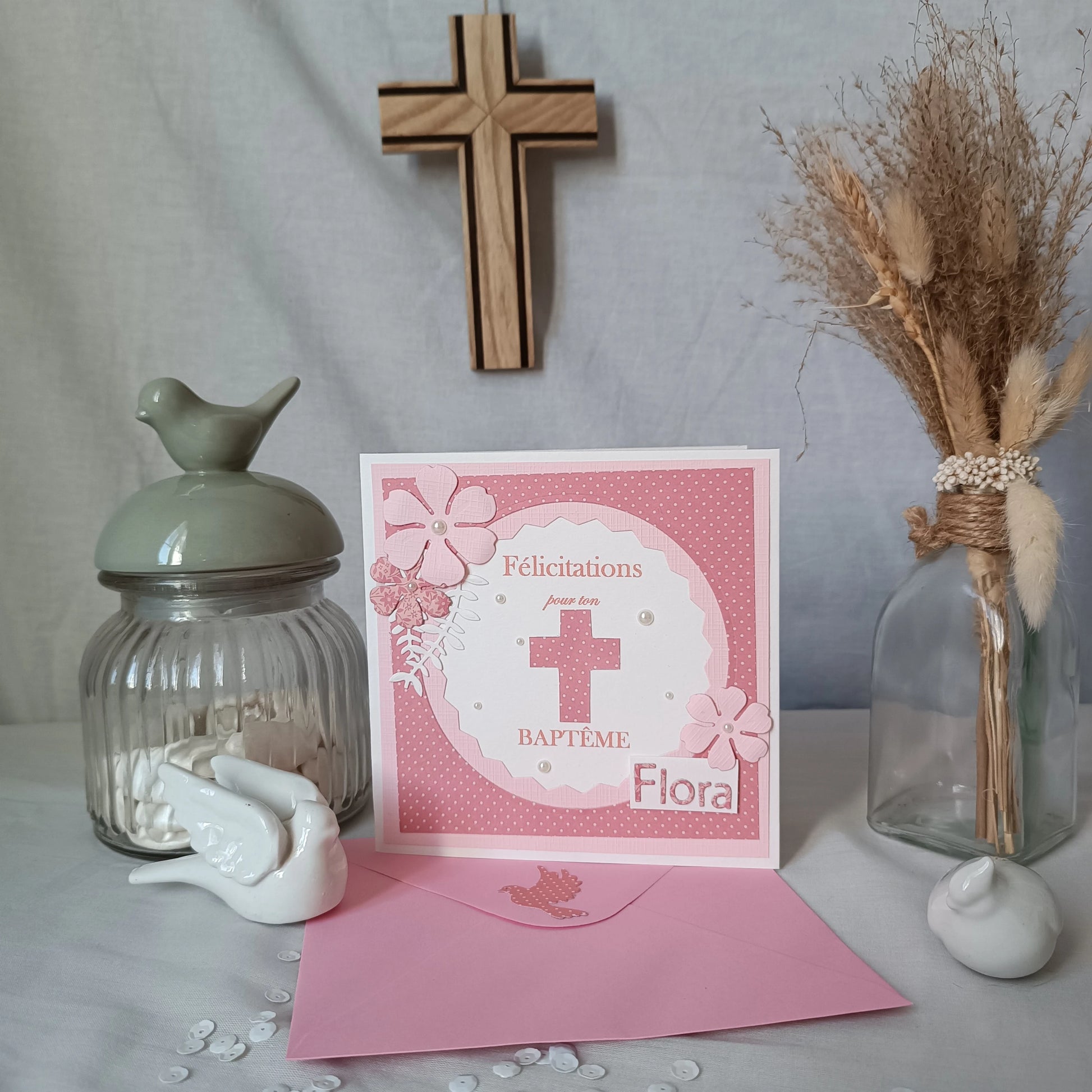 Carte de baptême rose personnalisable, debout sur son enveloppe rose, avec croix en bois et pot décoratif en arrière-plan