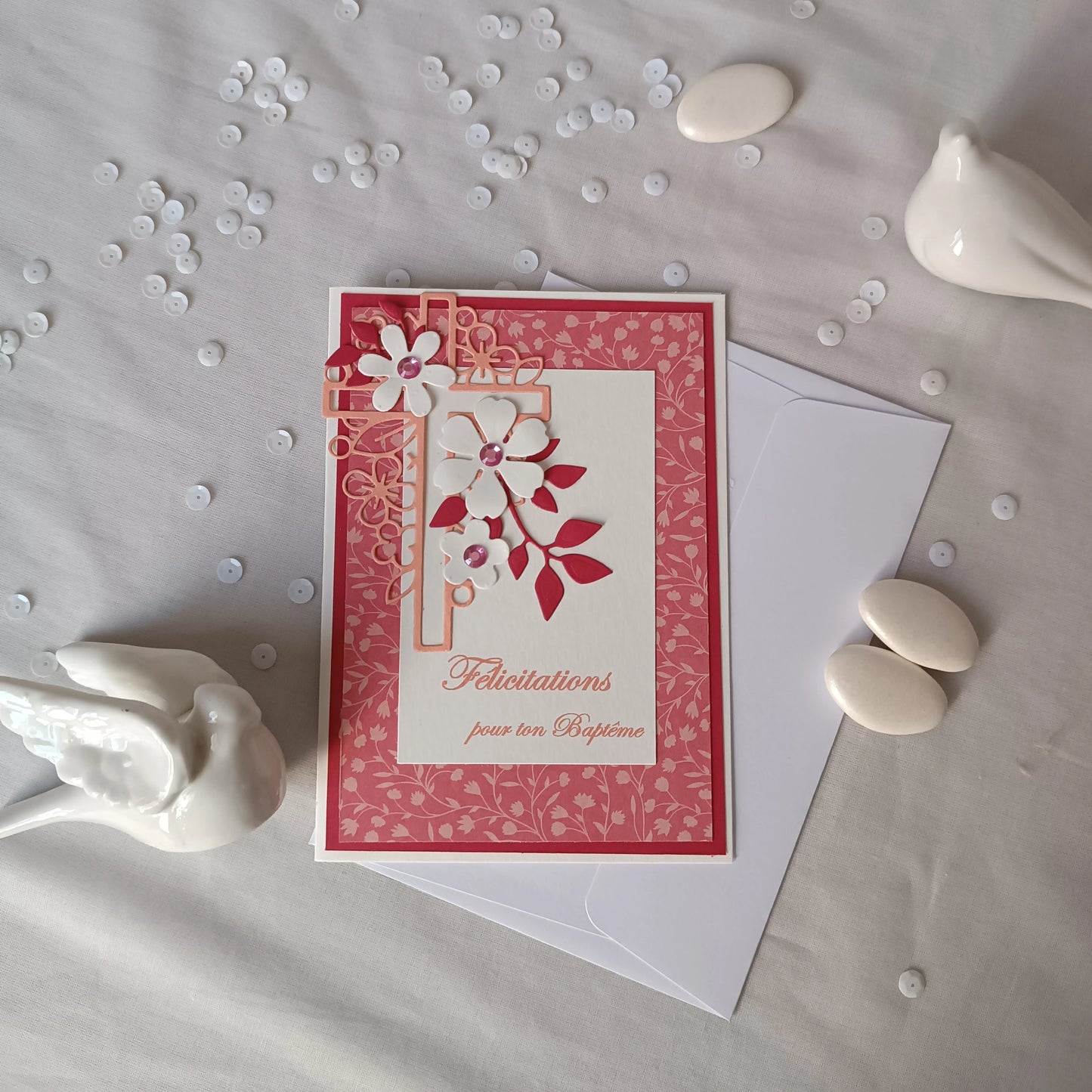 Vue rapproché de la carte de baptême rose posée sur un tissu, avec dragées et confettis décoratifs