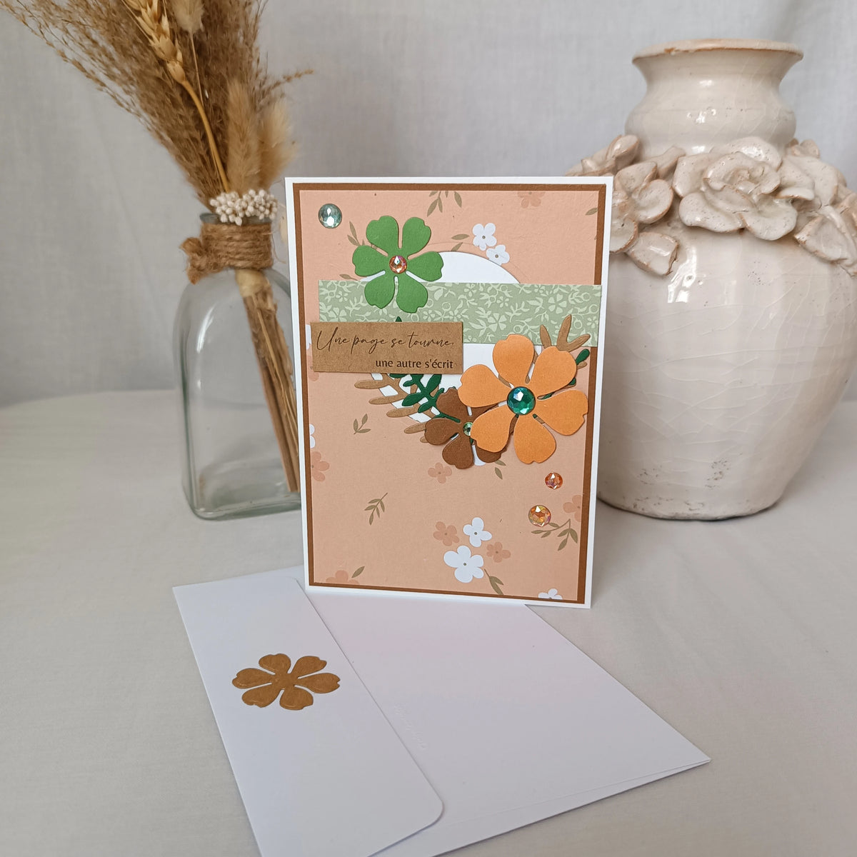 Carte de départ collègue abricot et vert sauge debout sur son enveloppe, entourée d'un vase de fleurs séchées et d'un pot en céramique