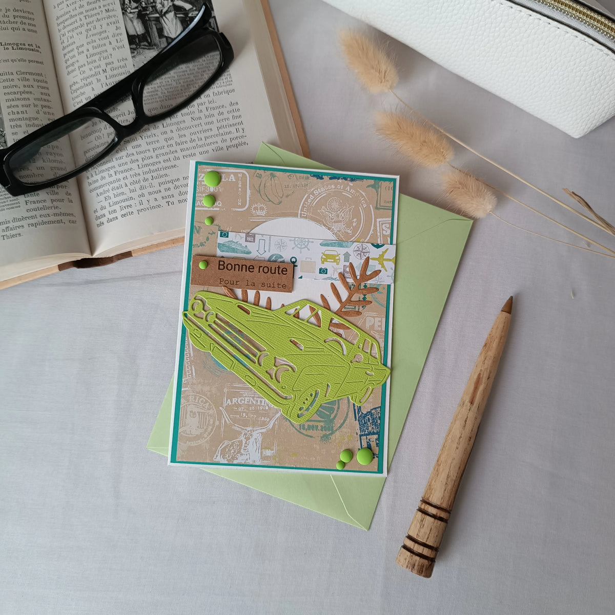 Vue en plongée de la carte de départ homme voiture posée sur son enveloppe, entourée d'une trousse, d'un livre ancien et de lunettes