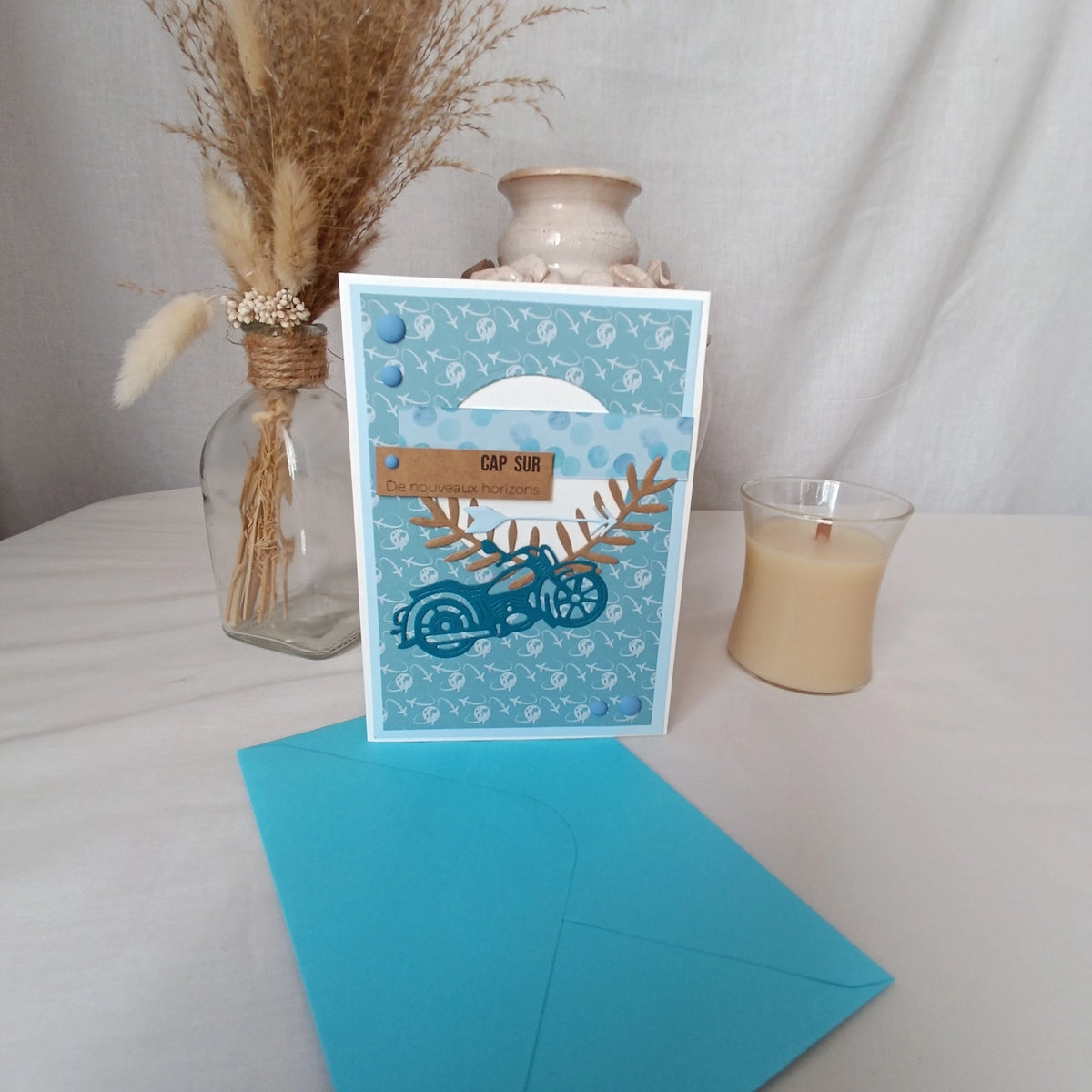 Carte de départ homme moto bleu sarcelle tenue debout devant un vase en céramique et une bougie, ambiance cosy