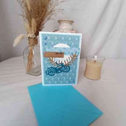 Carte de départ homme moto bleu sarcelle tenue debout devant un vase en céramique et une bougie, ambiance cosy