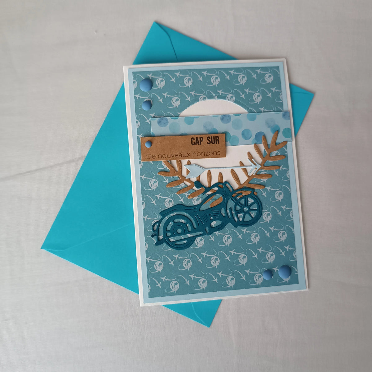 Carte de départ homme artisanale L'Atelier de Lili la souris, moto bleu sarcelle et motif avions, posée sur son enveloppe turquoise assortie