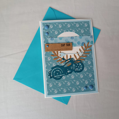 Carte de départ homme artisanale L'Atelier de Lili la souris, moto bleu sarcelle et motif avions, posée sur son enveloppe turquoise assortie