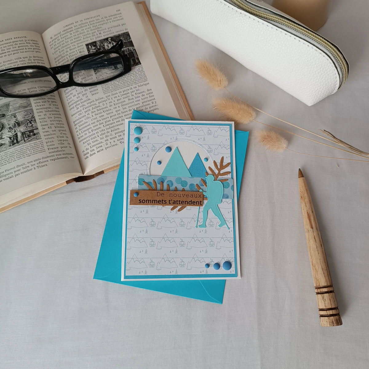 Carte de départ homme turquoise posée sur son enveloppe, entourée d'un livre ancien, de lunettes et d'un stylo en bois