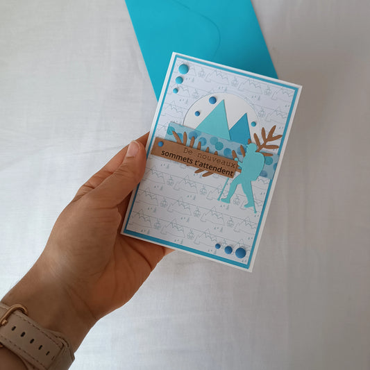 Carte de départ homme turquoise tenue à la main, montrant le randonneur et les montagnes découpés