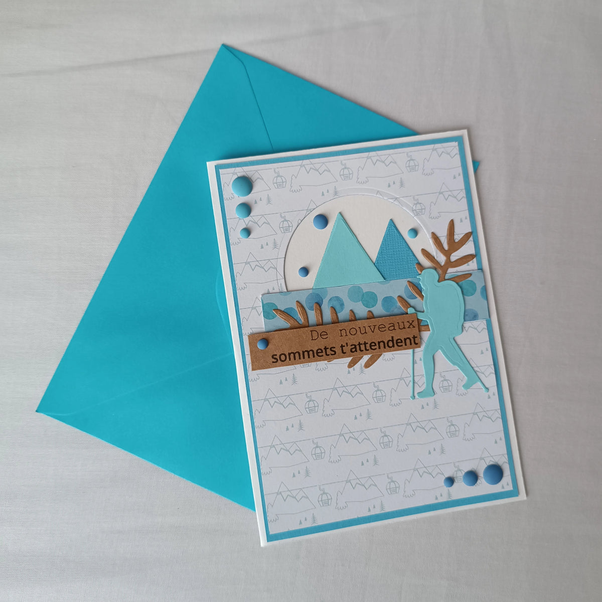 Carte de départ homme artisanale L'Atelier de Lili la souris, montagnes turquoise et randonneur, posée sur son enveloppe turquoise assortie