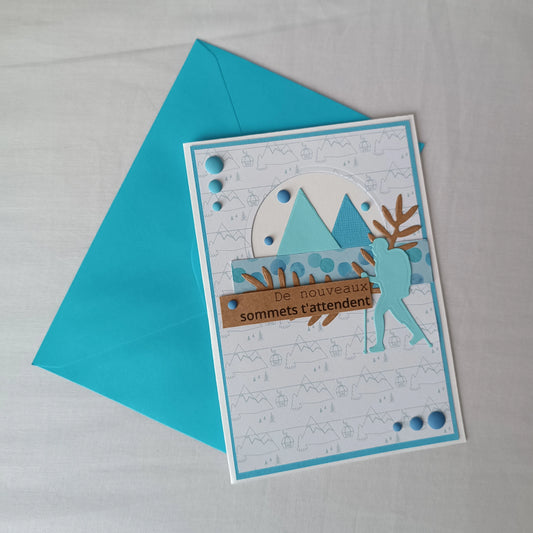 Carte de départ homme artisanale L'Atelier de Lili la souris, montagnes turquoise et randonneur, posée sur son enveloppe turquoise assortie